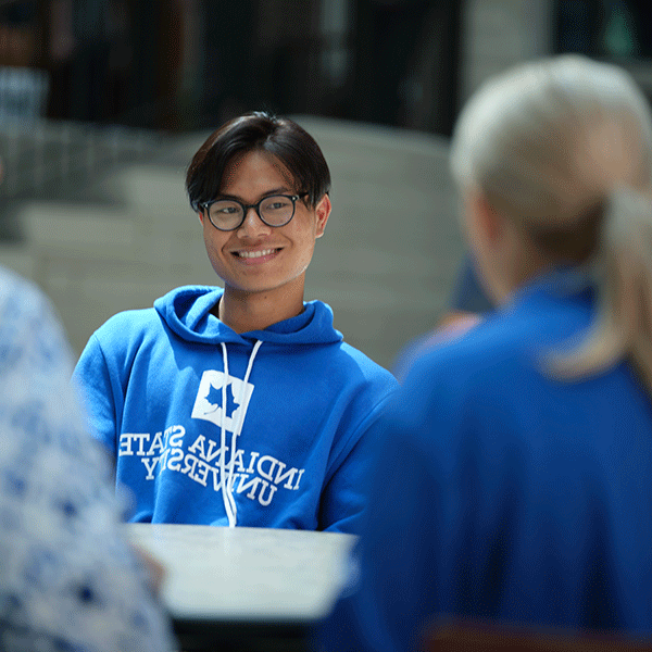 A young Asian American man with black hair and glasses smiles and chats with a blonde woman sitting opposite him. He is wearing a blue and white Indiana State University hoodie.